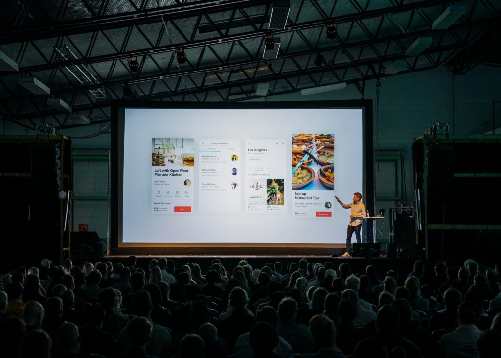 A person presents on a bright screen to a crowded room