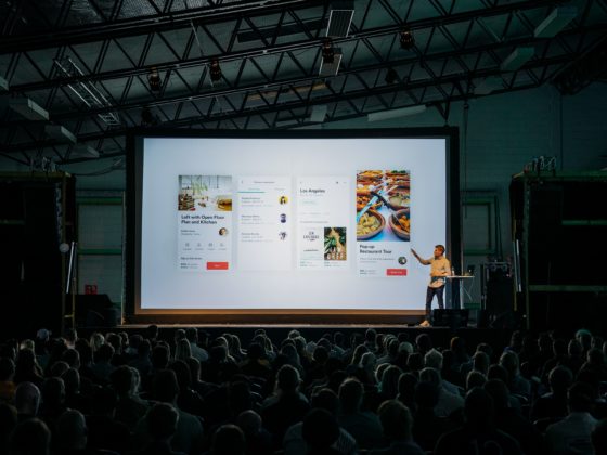 A person presents on a bright screen to a crowded room